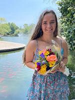 White woman with long dark blonde haor holding flowers in front of river