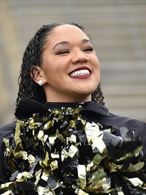 Smiling young lady with brown skin wearing a black long sleeve jacket holding cheer pom poms. She has short black curly hair.